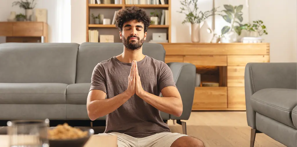 Mann sitzt meditierend im Wohnzimmer vor einem grauen Sofa und Holzmöbeln. Ruhige Wohnatmosphäre mit natürlichen Materialien, modernes Wohnen mit Komfort und stilvoller Einrichtung für mehr Entspannung im Alltag.